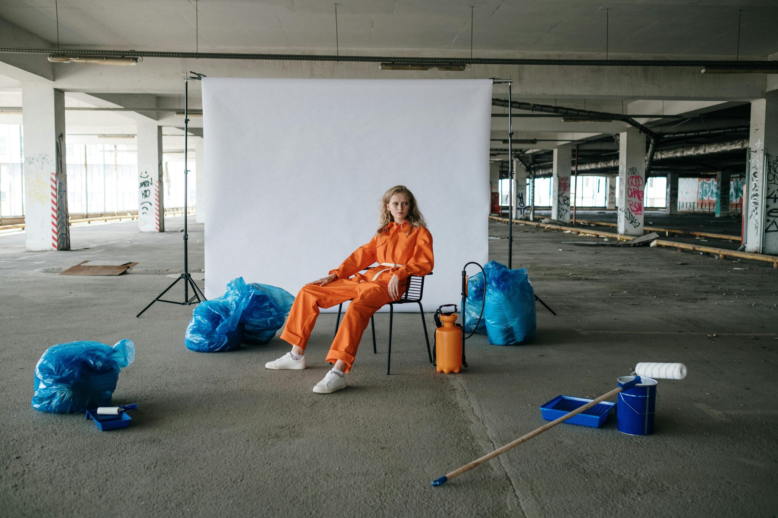 Artistic portrait of a woman in orange jumpsuit sitting in an industrial space with props.